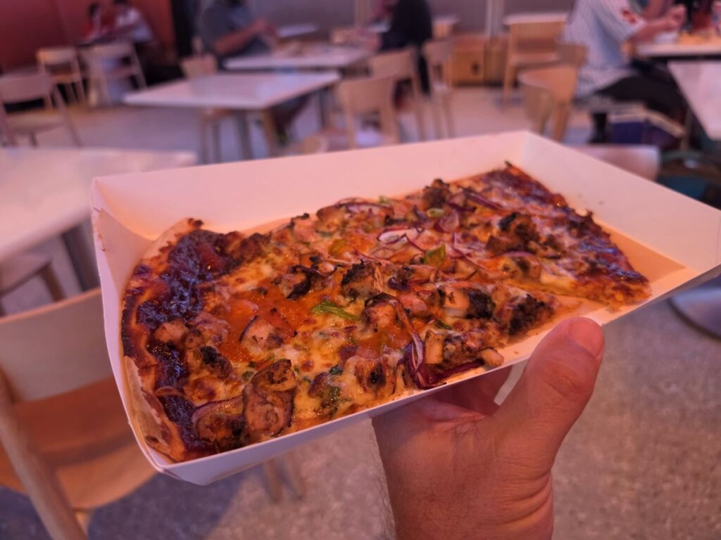 Hand holding a paper tray with two slices of BBQ Braai Chicken Pizza inside Connections Eatery at Epcot International Food & Wine Festival.