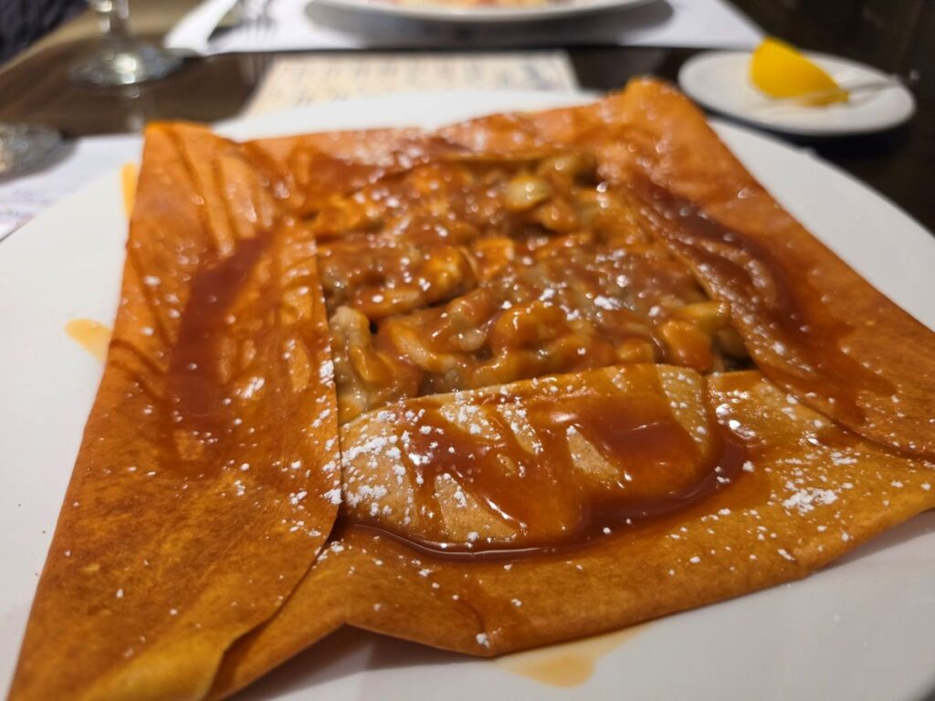 A golden-brown crepe folded on a white plate, topped with a rich caramel sauce and a light dusting of powdered sugar, served in a restaurant setting.