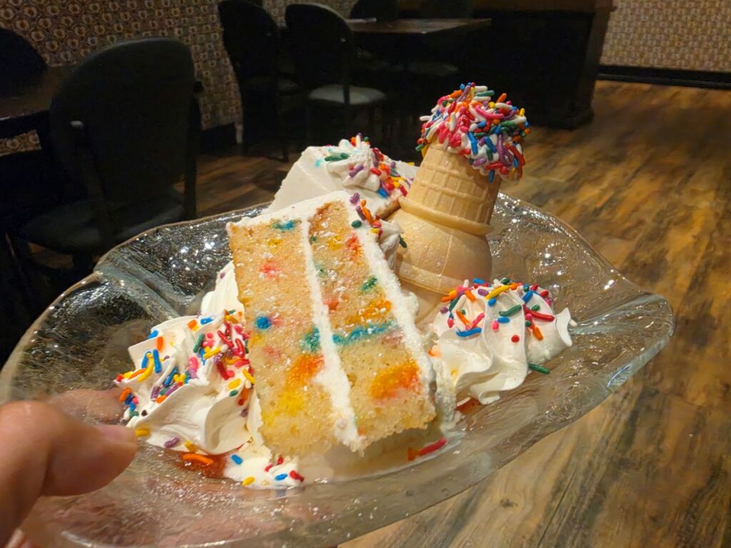 A lavish birthday cake sundae in a glass bowl featuring a slice of funfetti layer cake, an ice cream cone topped with sprinkles, and whipped cream at The Toothsome Chocolate Emporium.
