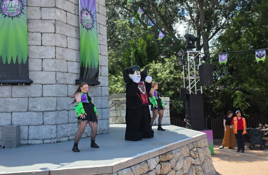 Performers in green and black costumes dancing alongside a Lego Lord Vampyre character on a stone stage during a live show.