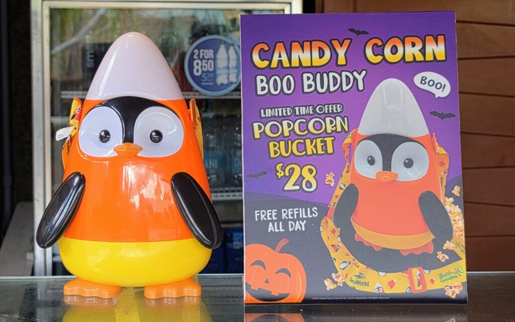 A penguin-shaped candy corn popcorn bucket stands next to a purple sign advertising the Candy Corn Boo Buddy bucket for twenty-eight dollars.