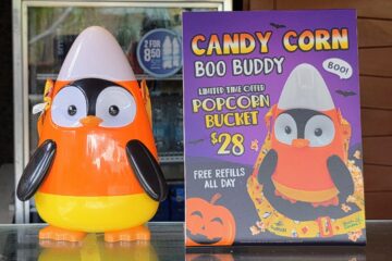 A penguin-shaped candy corn popcorn bucket stands next to a purple sign advertising the Candy Corn Boo Buddy bucket for twenty-eight dollars.