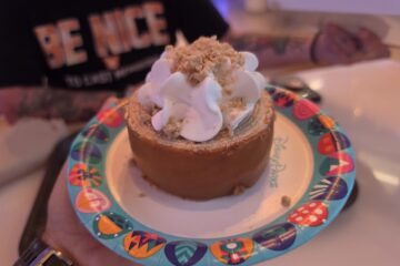 Person holding a Caramel-Apple Cheesecake Baumkuchen dessert topped with whipped cream inside Connections Eatery at Epcot.