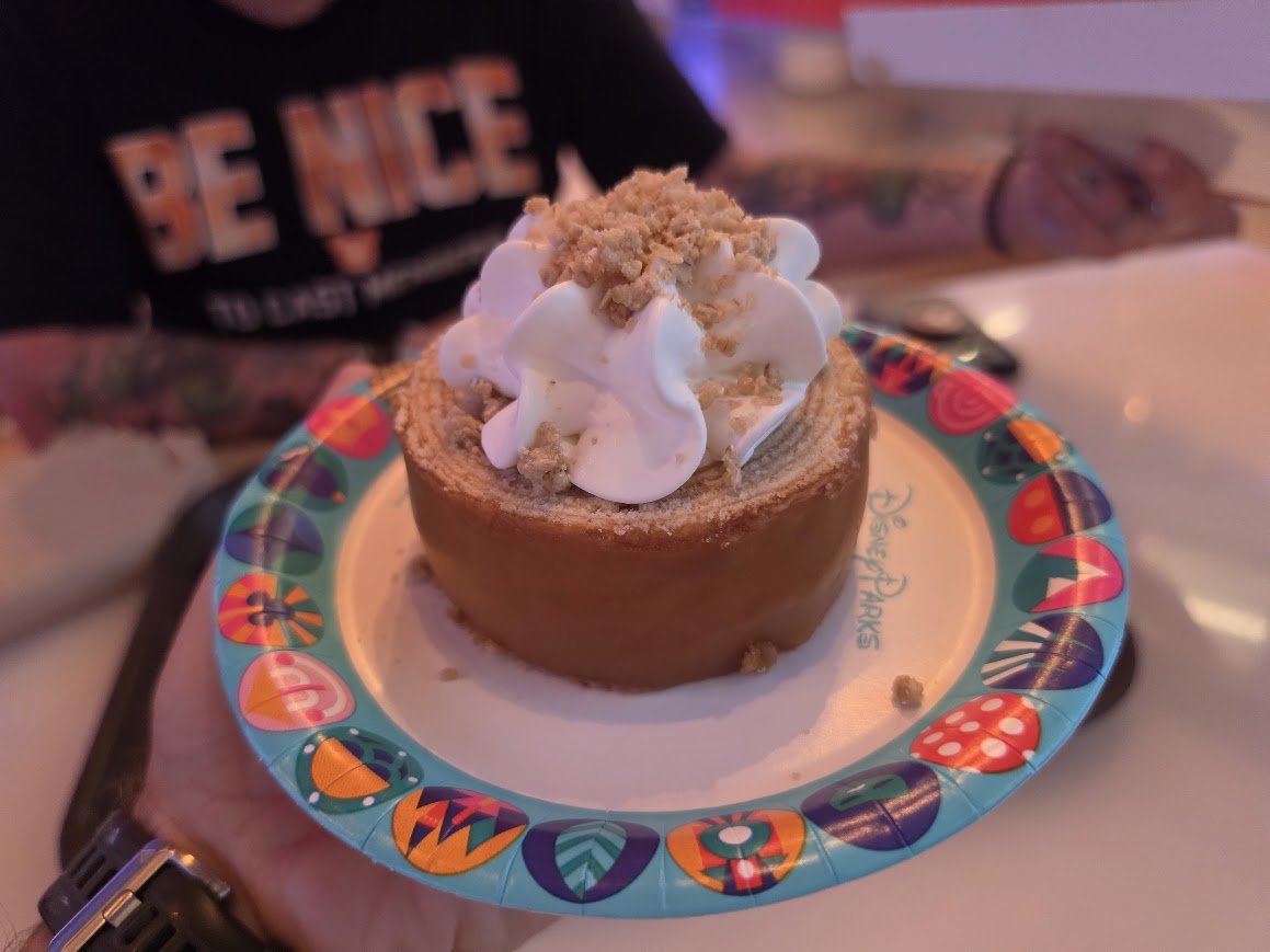 Person holding a Caramel-Apple Cheesecake Baumkuchen dessert topped with whipped cream inside Connections Eatery at Epcot.