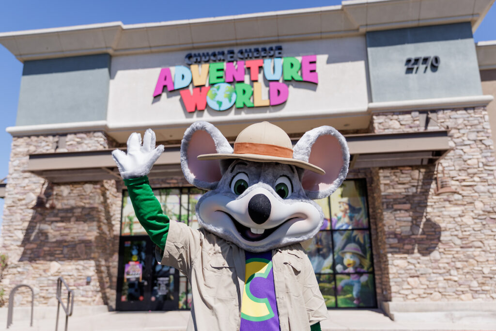 The Chuck E. Cheese mascot, wearing a safari hat and tan shirt, waves in front of a Chuck E. Cheese Adventure World building with stone exterior.
