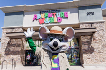 The Chuck E. Cheese mascot, wearing a safari hat and tan shirt, waves in front of a Chuck E. Cheese Adventure World building with stone exterior.