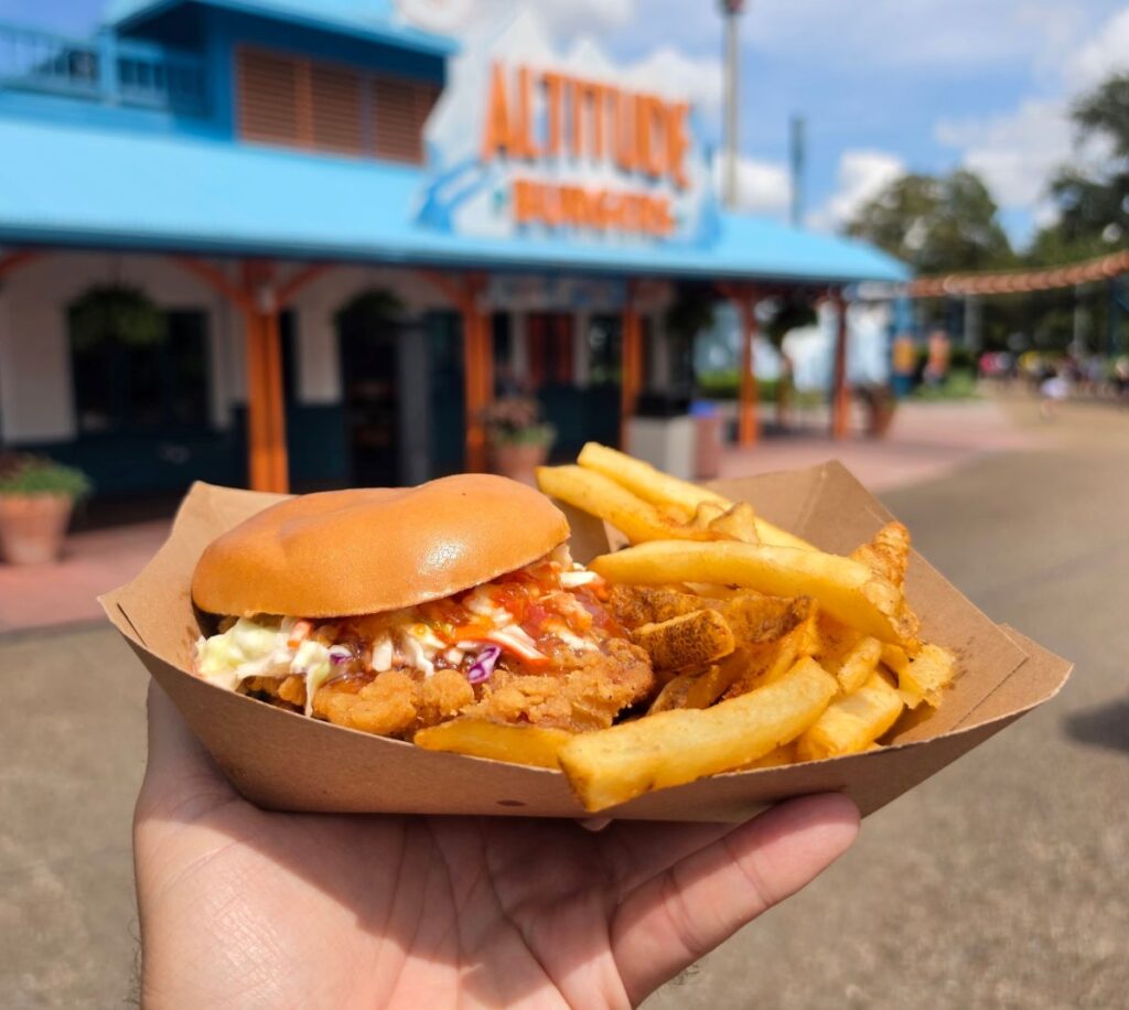 A hand holds a paper basket with a Crispy Thai chili chicken sandwich and French fries at Altitude Burgers in SeaWorld Orlando.