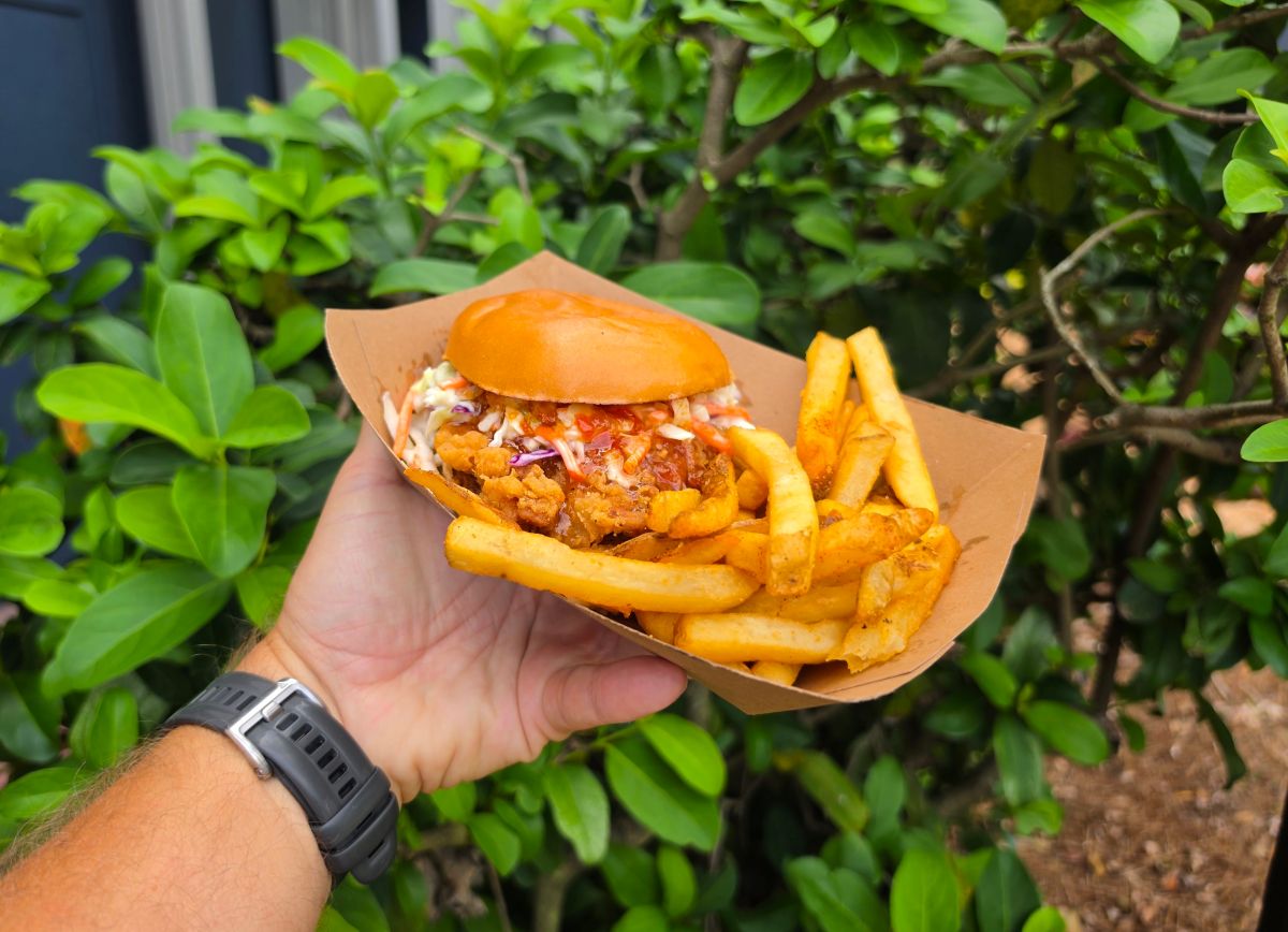 Hand holding a crispy Thai chili chicken sandwich and fries meal against leafy green bushes.