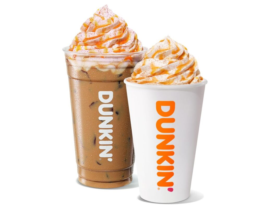 Two Dunkin' drinks: a clear plastic cup of iced coffee and a white paper cup of hot coffee, both topped with whipped cream and caramel drizzle.