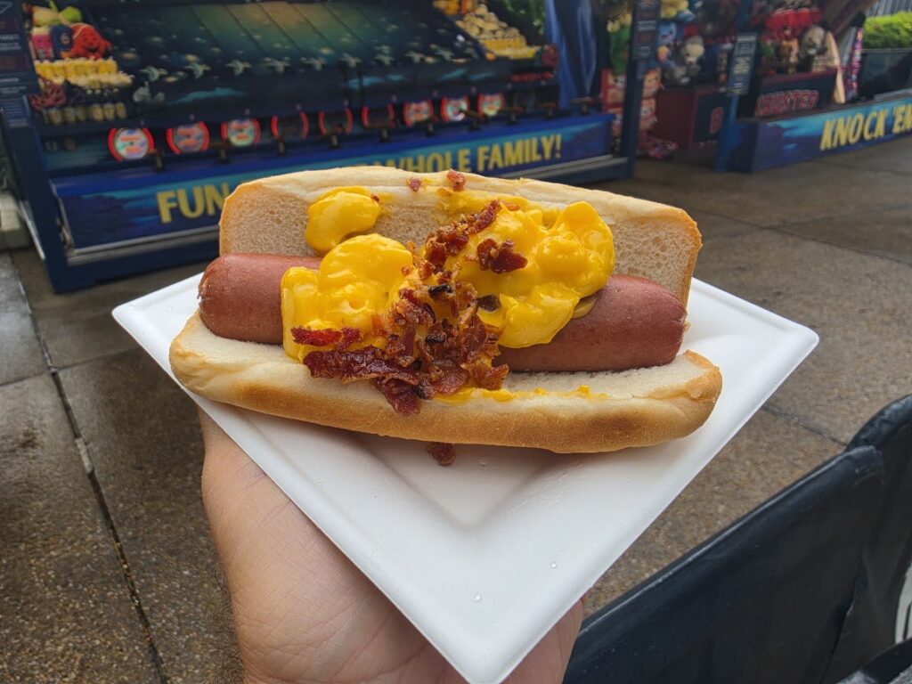 Highway Havoc Bacon Mac & Cheese Dog seen with a person holding it for Taste the Terror Menu at Howl-O-Scream Orlando