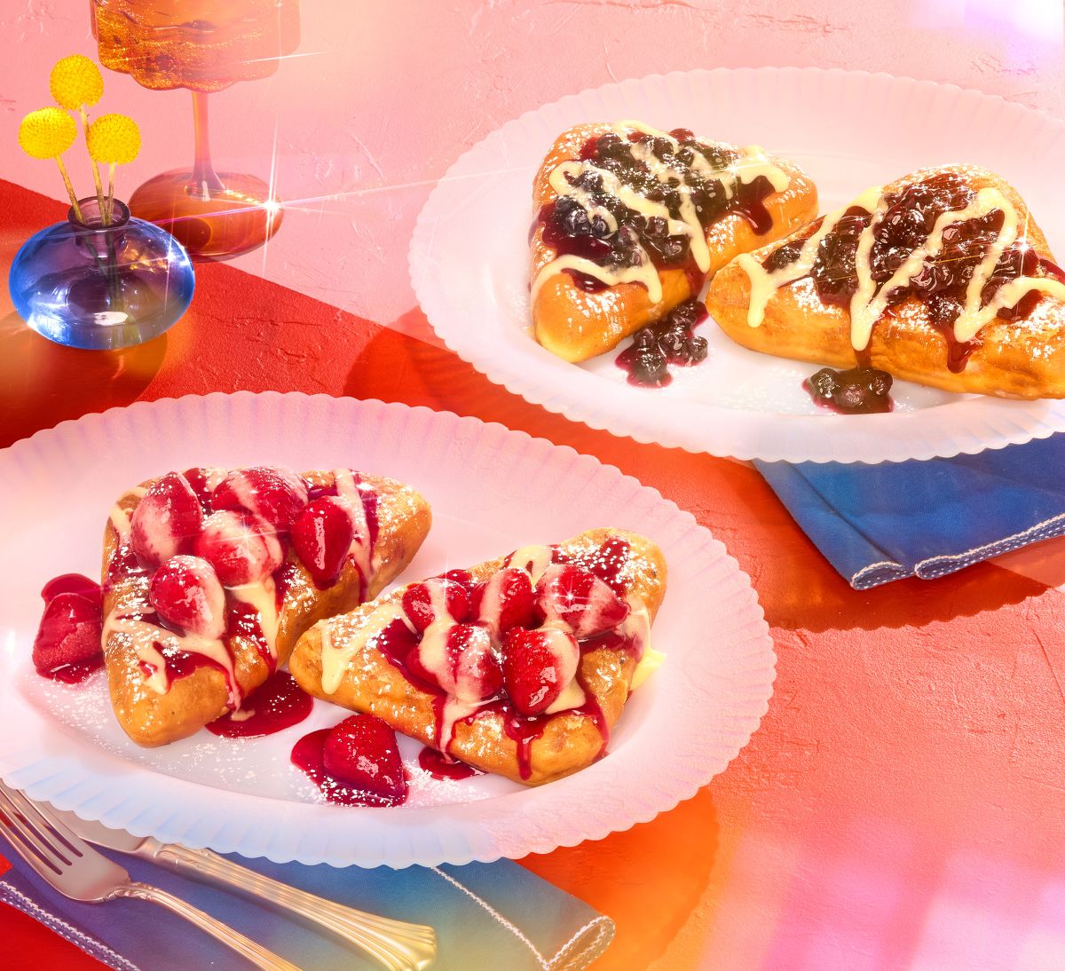 Two white plates hold IHOP Stuffed French Toast topped with strawberry or blueberry sauce and cream, set on a red surface with a blue napkin and flowers.