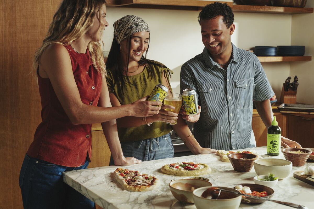 Three friends stand around a marble kitchen island, smiling and clinking cans of beverage over freshly prepared pizzas and bowls of ingredients.