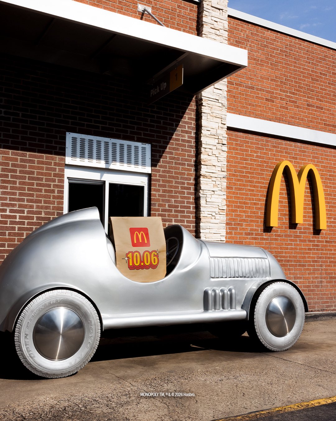 A vintage-style silver toy race car parked at a McDonald's drive-thru window with a brown paper bag featuring the McDonald's logo and the text 10.06 promoting McDonald's Monopoly game