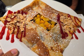 A square-folded crepe on a white plate, filled with diced yellow mango, topped with sliced almonds, red berry sauce, and a dusting of sugar.