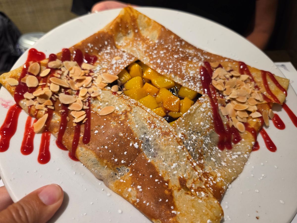 A square-folded crepe on a white plate, filled with diced yellow mango, topped with sliced almonds, red berry sauce, and a dusting of sugar.