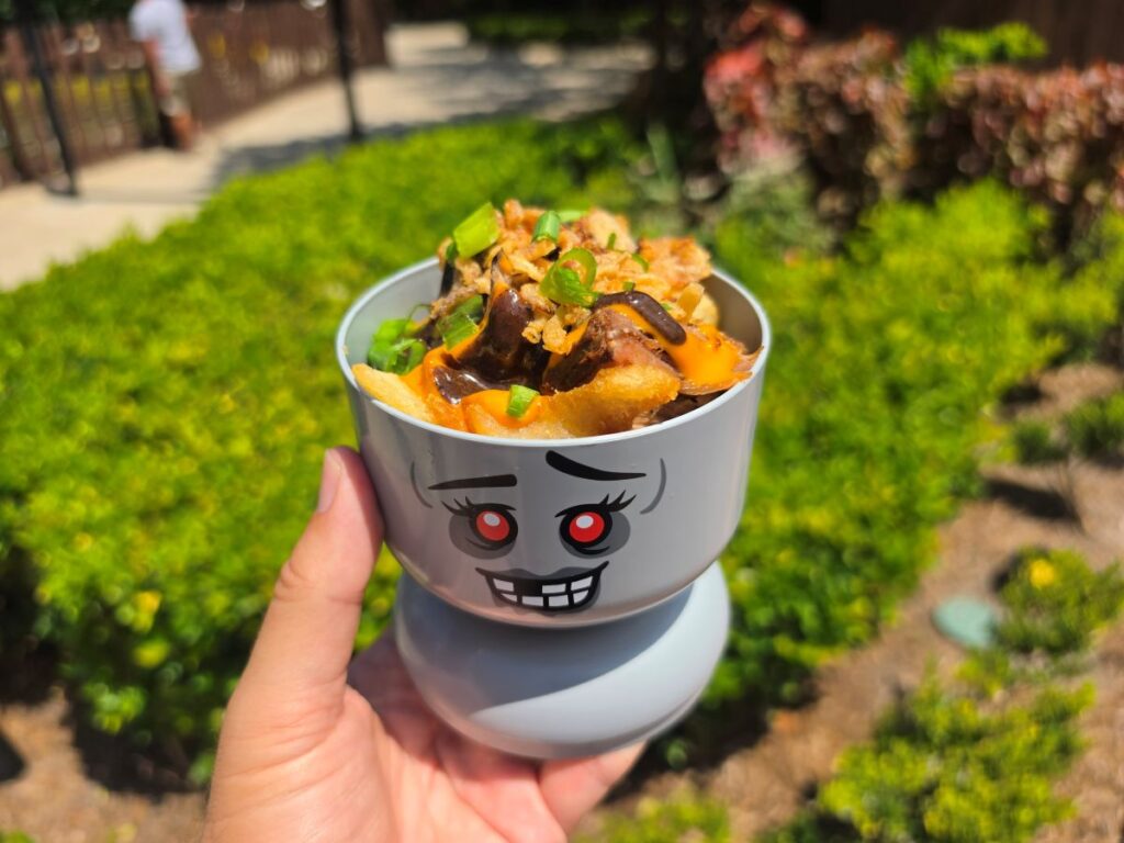 Hand holding a gray Lego monster-faced dish filled with loaded fries, cheese sauce, and green onions outdoors for Brick or Treat at Legoland Florida Resort