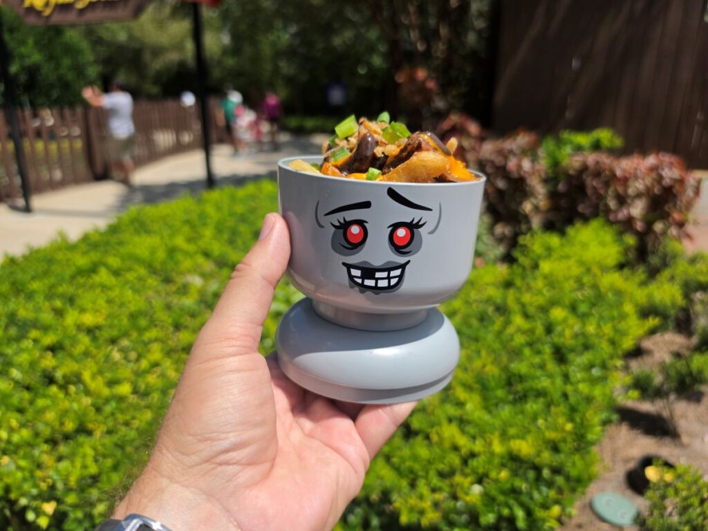 Close-up view of hand holding a gray Lego monster cup loaded with fries, cheese, melted sauce, and toppings against a garden backdrop.
