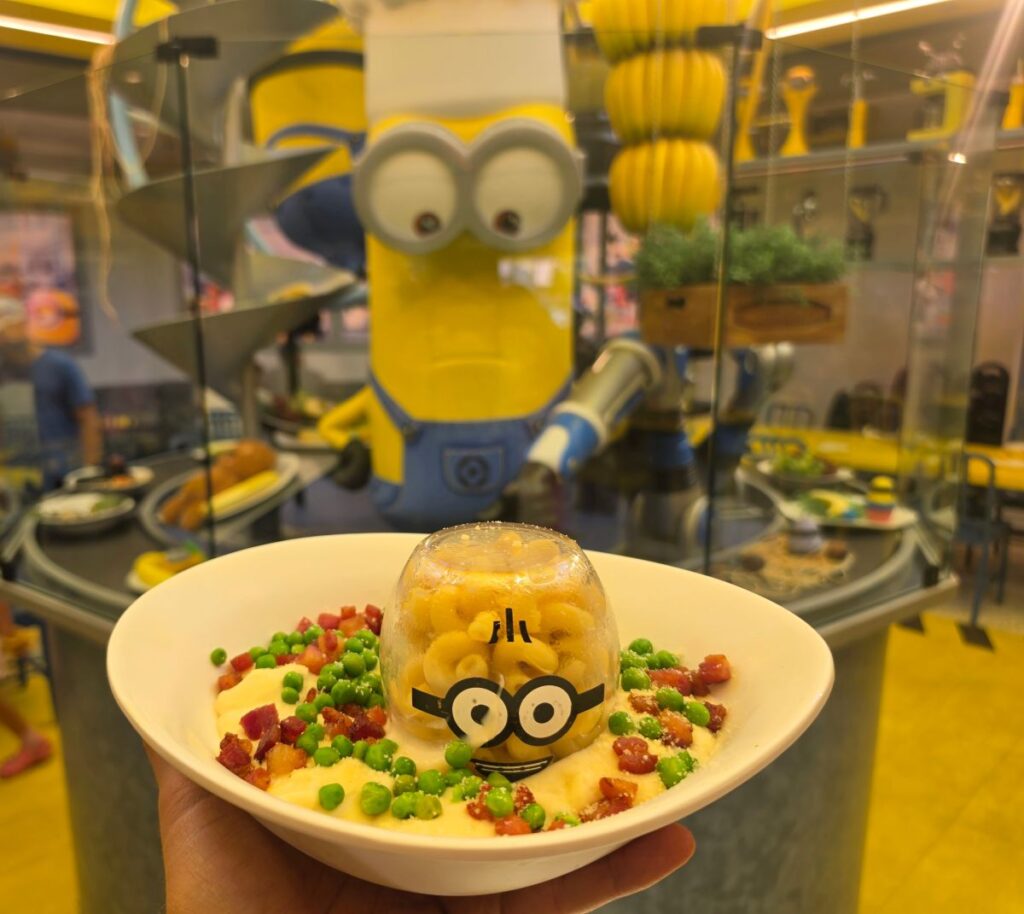 A hand holds a bowl of Otto’s Carbonara featuring a clear dome with a Minion face, in front of a large Minion chef statue.