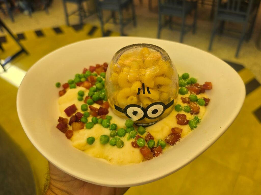 A white bowl holds Otto’s Carbonara with green peas and bacon bits, and a clear glass dome holds macaroni with a Minion face design.