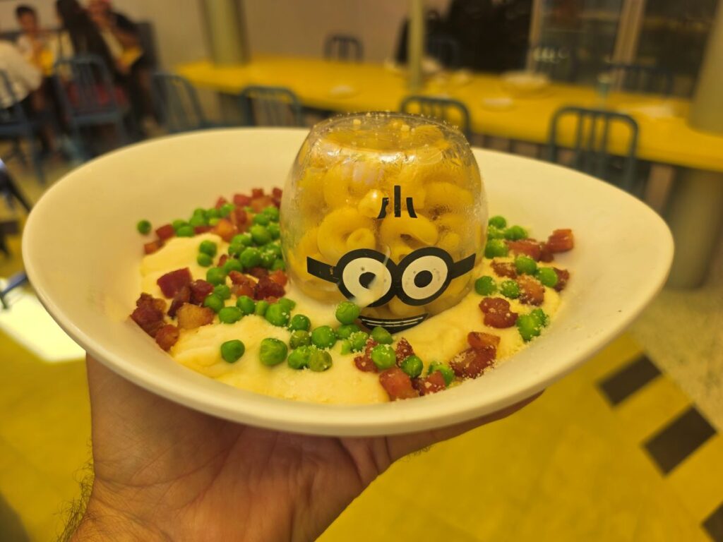 A white bowl holds a cream sauce topped with green peas and bacon bits, and a clear glass dome holds macaroni with a Minion face design.