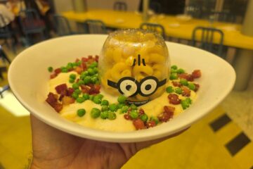 A white bowl holds a cream sauce topped with green peas and bacon bits, and a clear glass dome holds macaroni with a Minion face design.