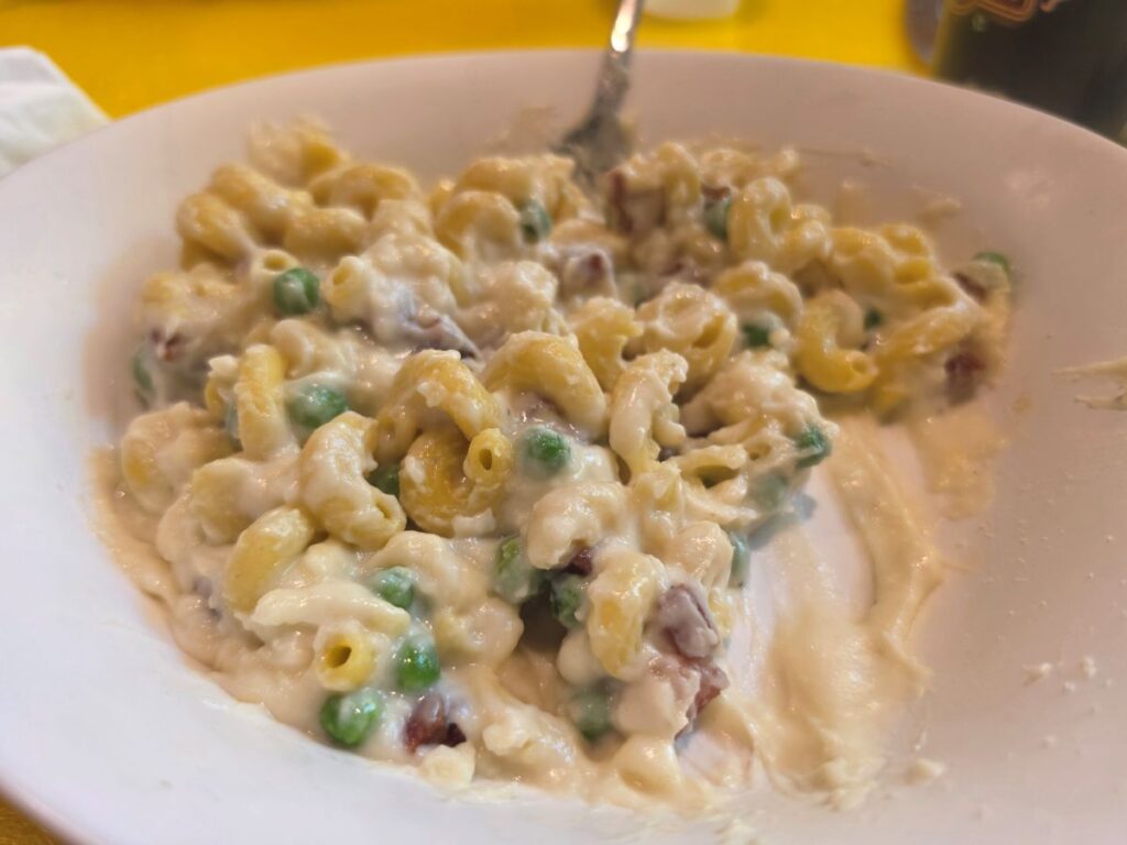 A white bowl filled with Otto’s Carbonara with mixed pasta mixed, green peas and small pieces of pancetta, served against a bright yellow background surface.