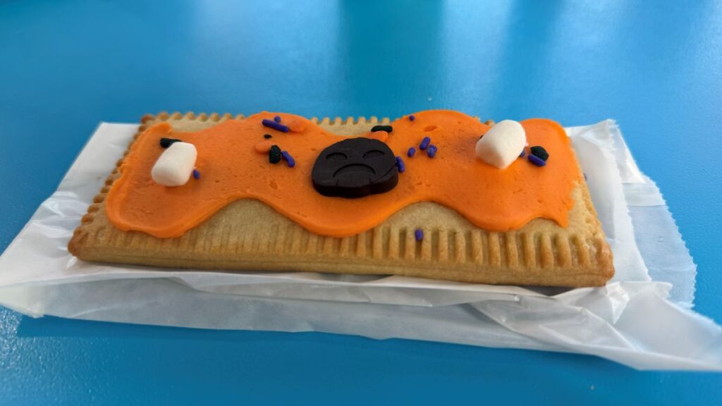 A rectangular Pumpkin Lunch Box Tart toaster pastry with orange frosting, two small marshmallows, purple sprinkles, and a dark chocolate piece shaped like a sad face.