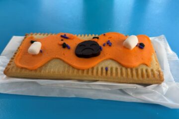 A rectangular toaster pastry with orange frosting, two small marshmallows, purple sprinkles, and a dark chocolate piece shaped like a sad face.