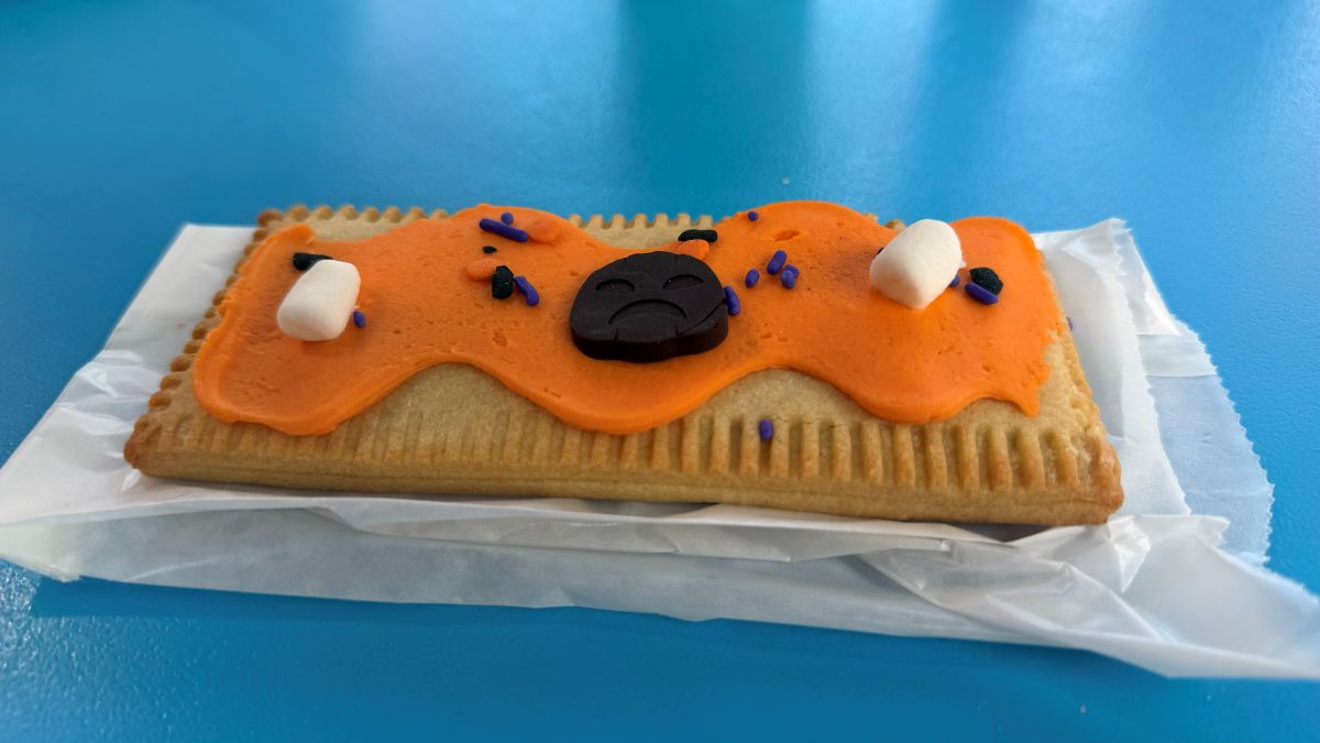A rectangular toaster pastry with orange frosting, two small marshmallows, purple sprinkles, and a dark chocolate piece shaped like a sad face.