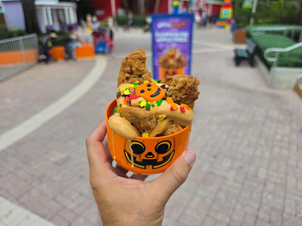 Hand holding a Pumpkin Pie Apple Fries jack-o'-lantern cup packed with warm apple fries, soft-serve whip, cookie pieces, and leaf-shaped sprinkles.