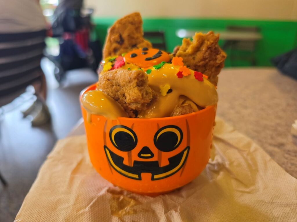 Close-up of a Pumpkin Pie Apple Fries jack-o'-lantern bowl filled with soft-serve topped apple fries, cookies, and colorful autumn sprinkles on a table.
