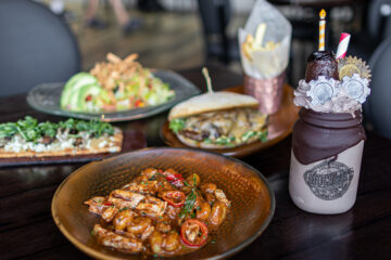 A variety of dishes on a wooden table, including shrimp in sauce, a sandwich, a salad, and a decorative milkshake with toppings.