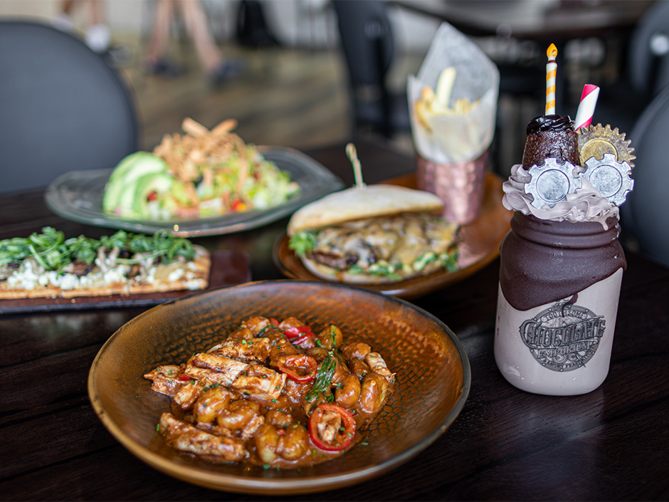 A variety of dishes on a wooden table, including shrimp in sauce, a sandwich, a salad, and a decorative milkshake with toppings.