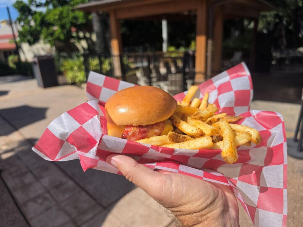 A hand holds a basket lined with red and white checkered paper containing a cheeseburger with bacon and a side of seasoned golden french fries.