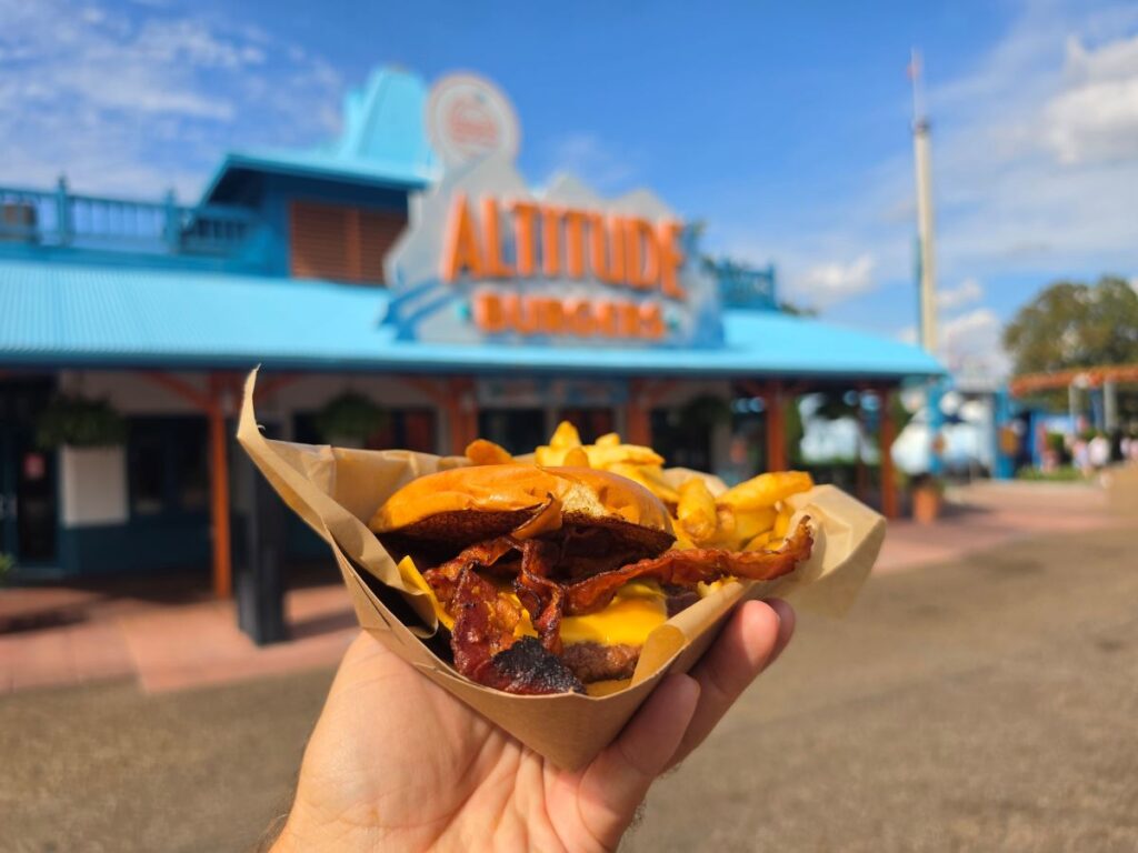 A hand holds a paper tray with a bacon cheeseburger and fries in front of the blue Altitude Burgers restaurant building on a sunny, clear day.