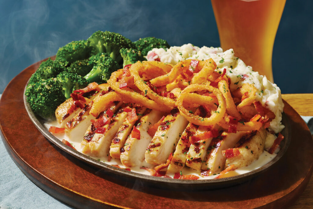 A sizzling platter features sliced grilled chicken topped with bacon and onion rings, served with steamed broccoli and creamy mashed potatoes nearby.