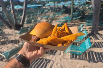 A hand holds a cardboard tray with a cheeseburger and french fries at a beach resort, with rows of empty teal lounge chairs in the blurred background.