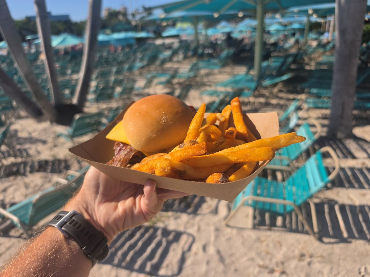 A hand holds a cardboard tray with a cheeseburger and french fries at a beach resort, with rows of empty teal lounge chairs in the blurred background.