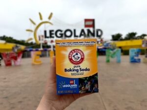 A hand holds an Arm & Hammer baking soda box featuring a Legoland promotional offer in front of the blurred Legoland entrance sign and park area.