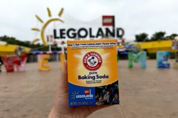 A hand holds an Arm & Hammer baking soda box featuring a Legoland promotional offer in front of the blurred Legoland entrance sign and park area.