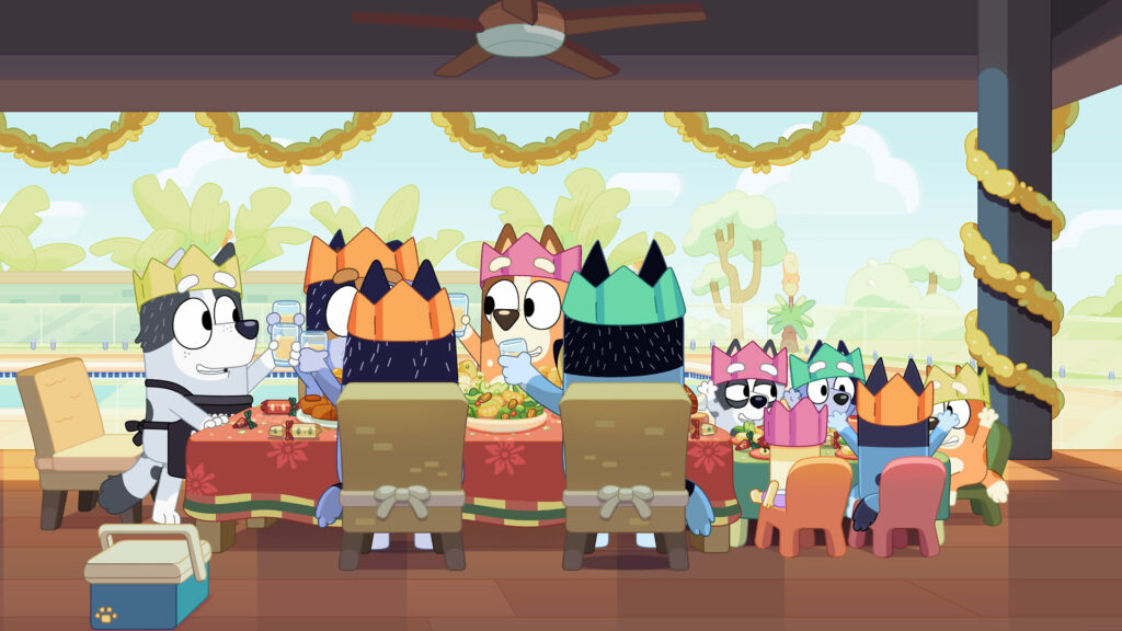 Cartoon dogs from Bluey wearing paper crowns sit around a festive dining table on a patio, raising glasses for a toast during a holiday meal.