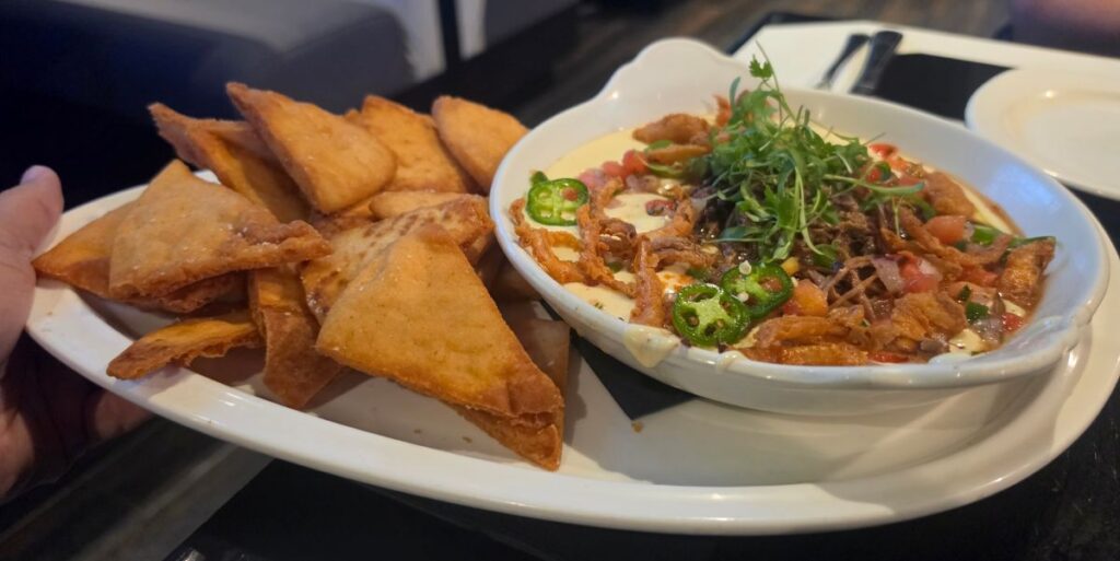 A white platter holds a bowl of creamy Brisket Queso & Chips dip topped with jalapeño slices, pico de gallo, and fresh herbs, served with crispy fried tortilla chips.