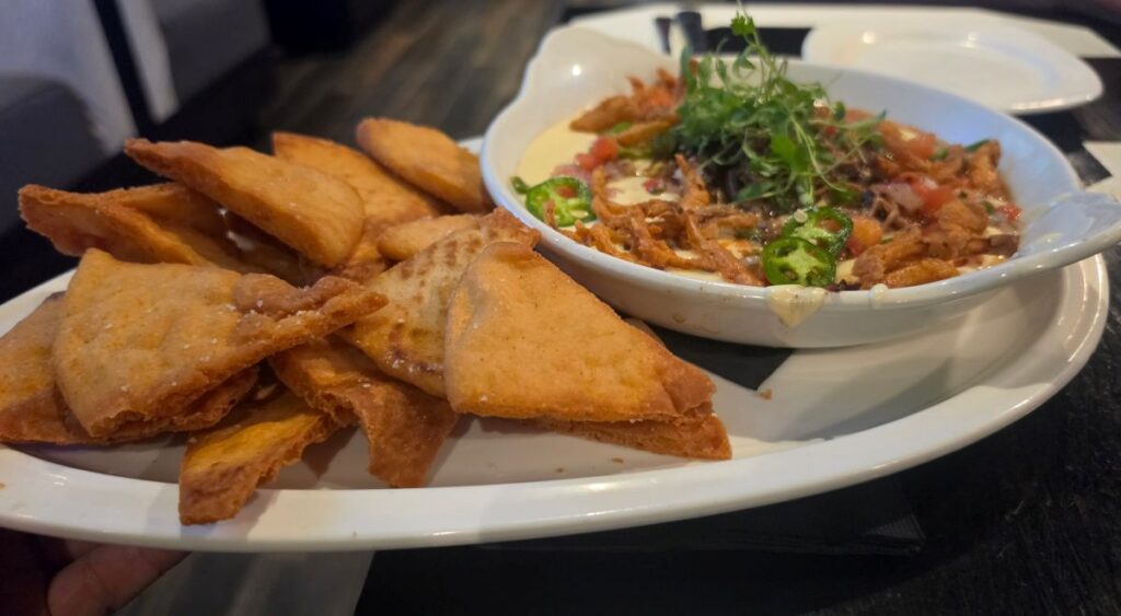 A white platter holds a bowl of creamy queso dip topped with fried onions, diced tomatoes, sliced jalapeños, and herbs, served with crispy chips.