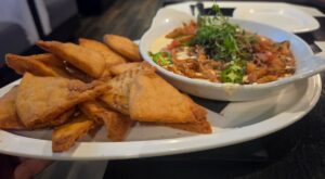 A white platter holds a bowl of creamy queso dip topped with fried onions, diced tomatoes, sliced jalapeños, and herbs, served with crispy chips.