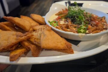 A white platter holds a bowl of creamy queso dip topped with fried onions, diced tomatoes, sliced jalapeños, and herbs, served with crispy chips.