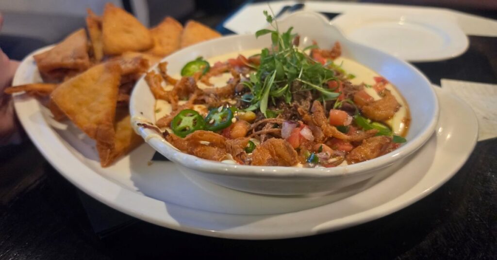 A white bowl of creamy dip topped with jalapeño slices, pico de gallo, and fresh herbs, served with a side of crispy fried tortilla triangles.