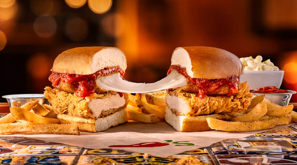 A chicken parmesan sandwich cut in half, showing melted cheese stretching between the two sides, served with seasoned fries on a patterned table.
