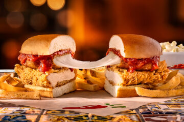 A chicken parmesan sandwich cut in half, showing melted cheese stretching between the two sides, served with seasoned fries on a patterned table.