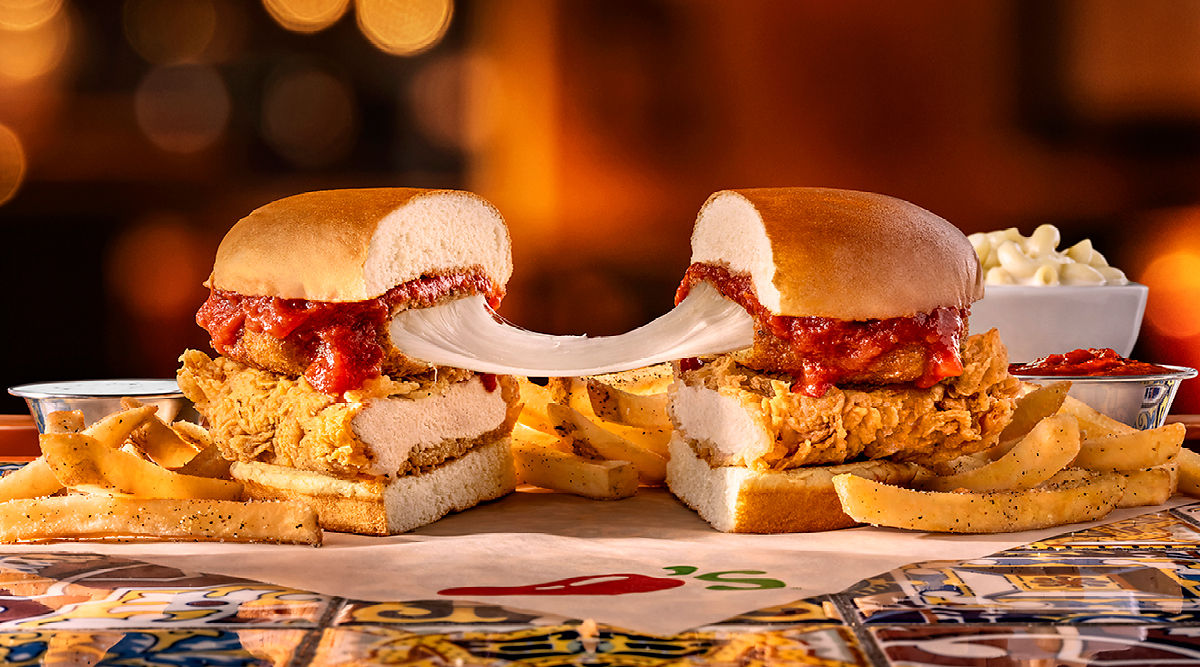 A chicken parmesan sandwich cut in half, showing melted cheese stretching between the two sides, served with seasoned fries on a patterned table.