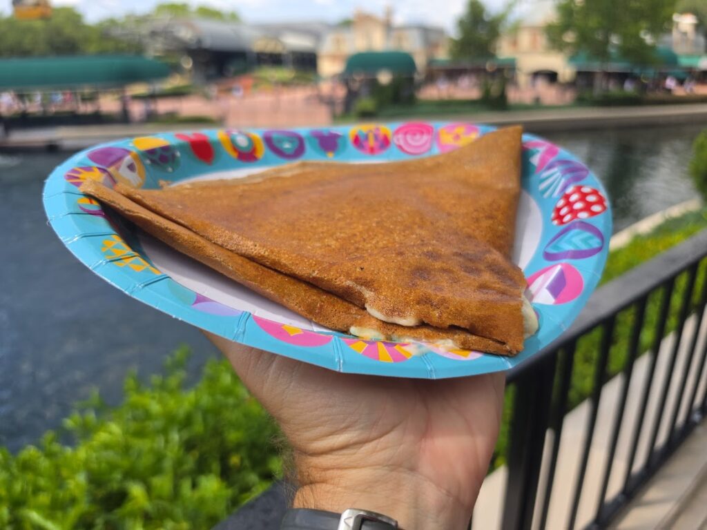 A hand holds a folded, golden-brown galette on a colorful paper plate with a patterned rim, set against a blurred park-and-water backdrop.